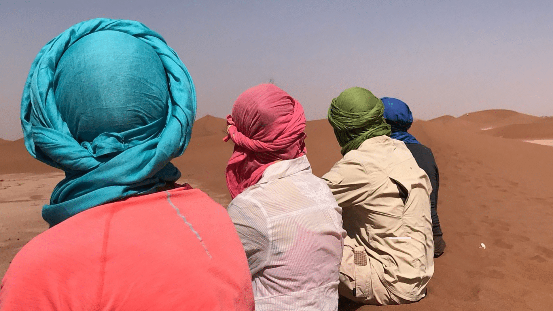 Un groupe de quatre personnes qui sont de dos méditent sur les dunes avec leur chèche.