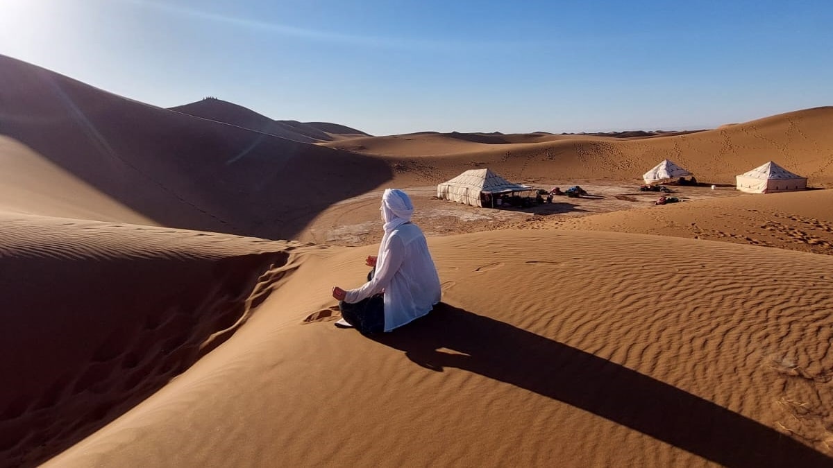 Une femme médite sur une dune, on aperçoit son bivouac avec les tentes au loin.