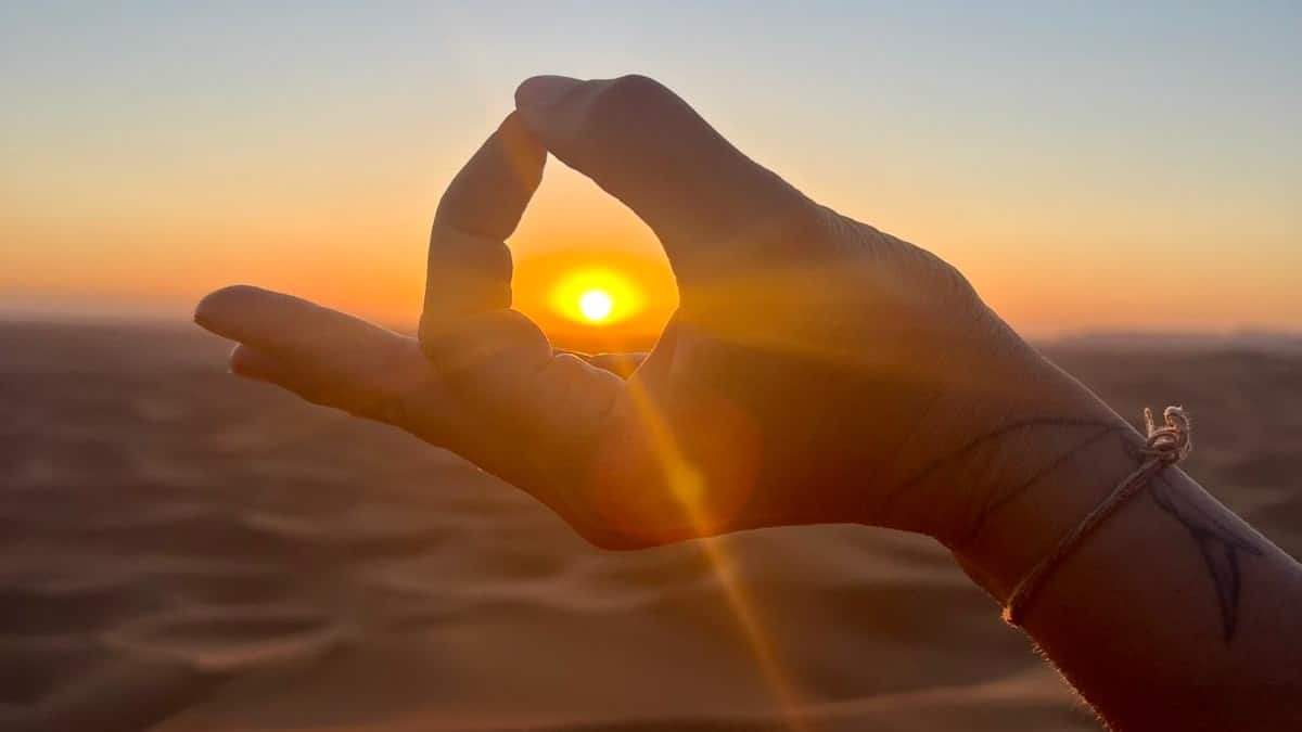 Une main en premier plan capture le soleil du désert en fond.